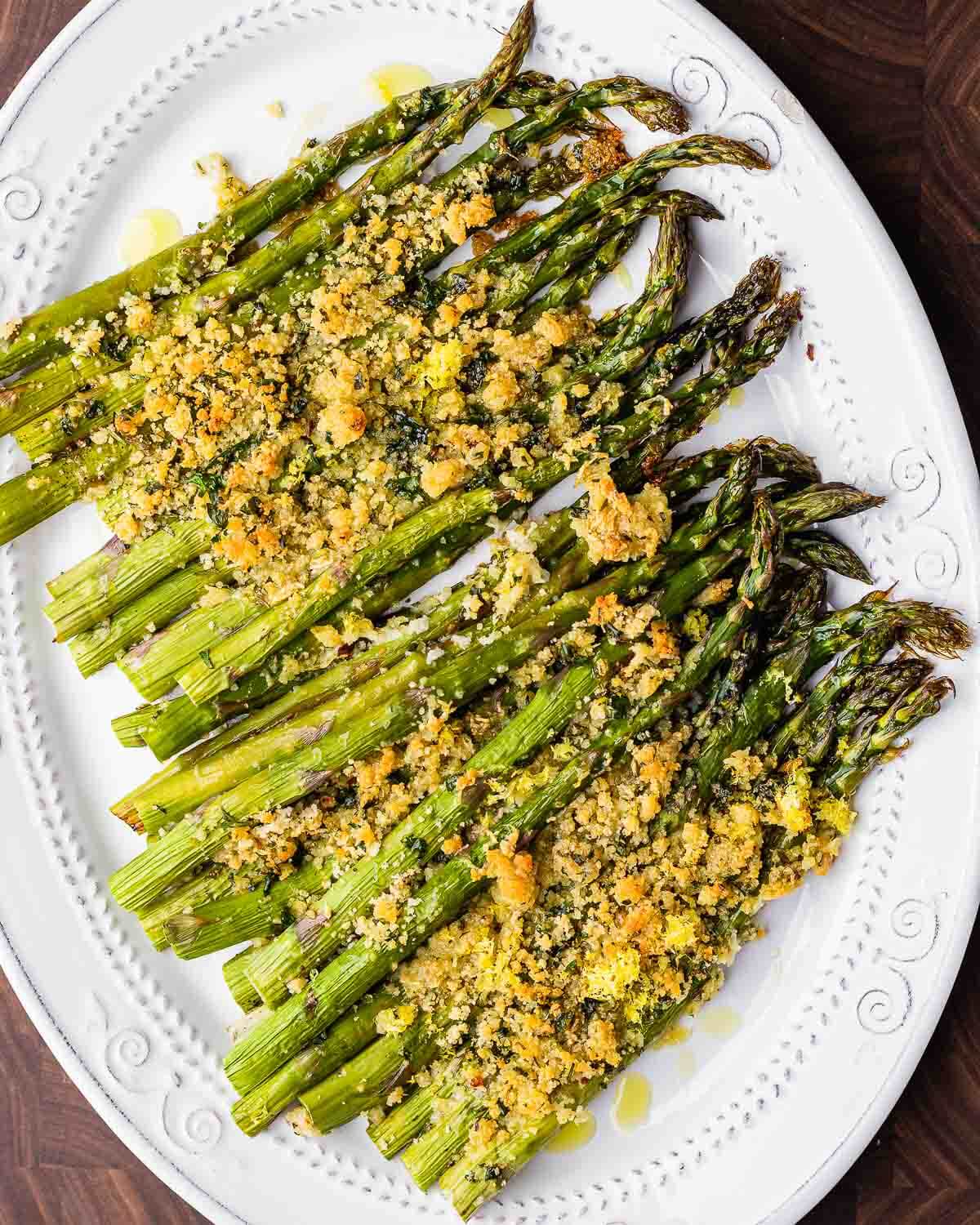 Overhead shot of baked asparagus with cheese and breadcrumbs on white platter.