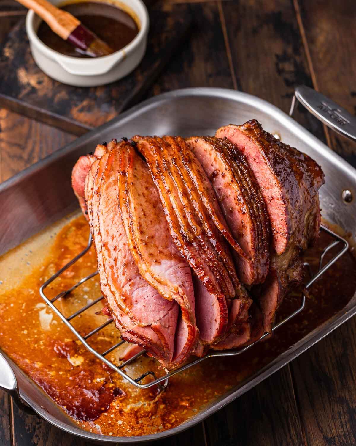 Brown sugar glazed spiral ham in roasting pan with bowl of extra glaze in the background.