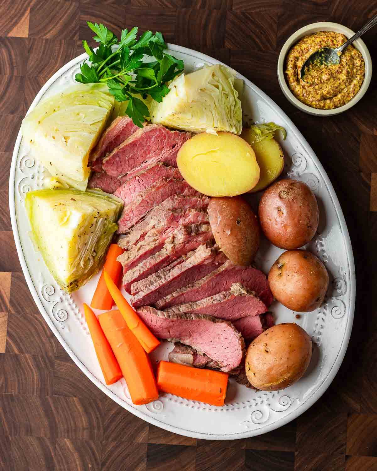 Overhead shot of platter with corned beef, cabbage, potatoes, and carrots along with small bowl of mustard on walnut cutting board.