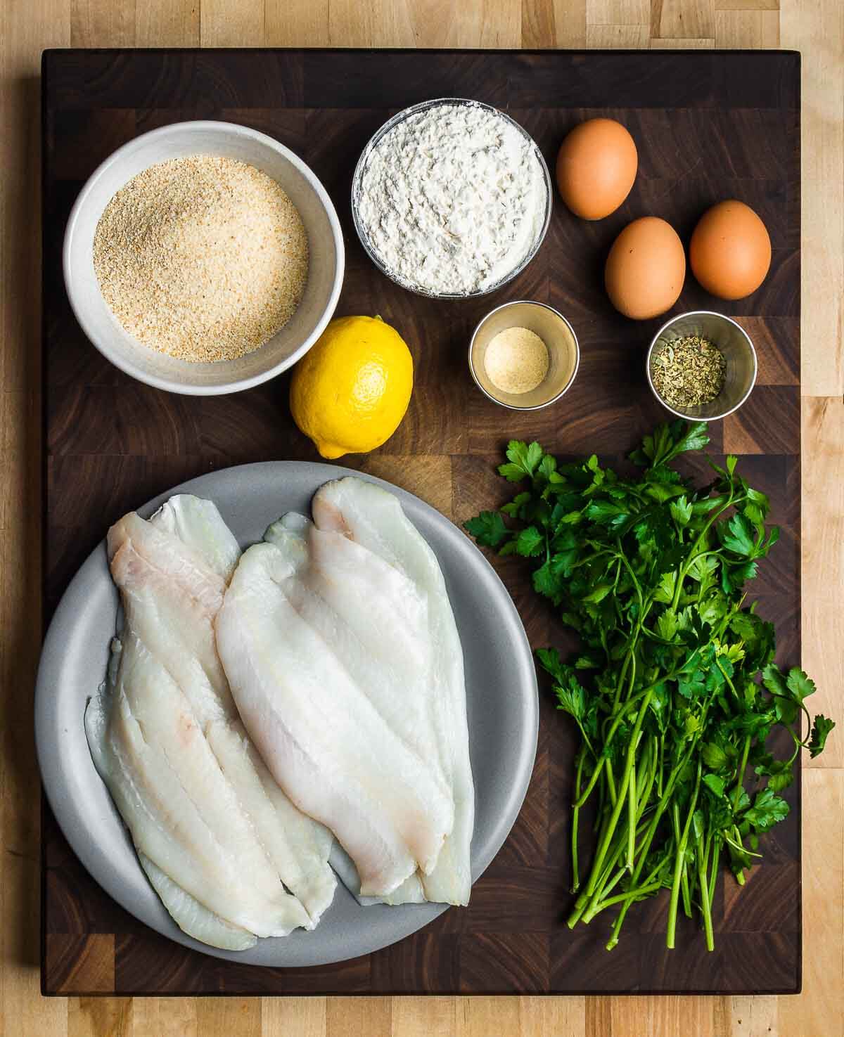 Ingredients shown: breadcrumbs, flour, lemon, garlic powder, eggs, oregano, flounder, and parsley.