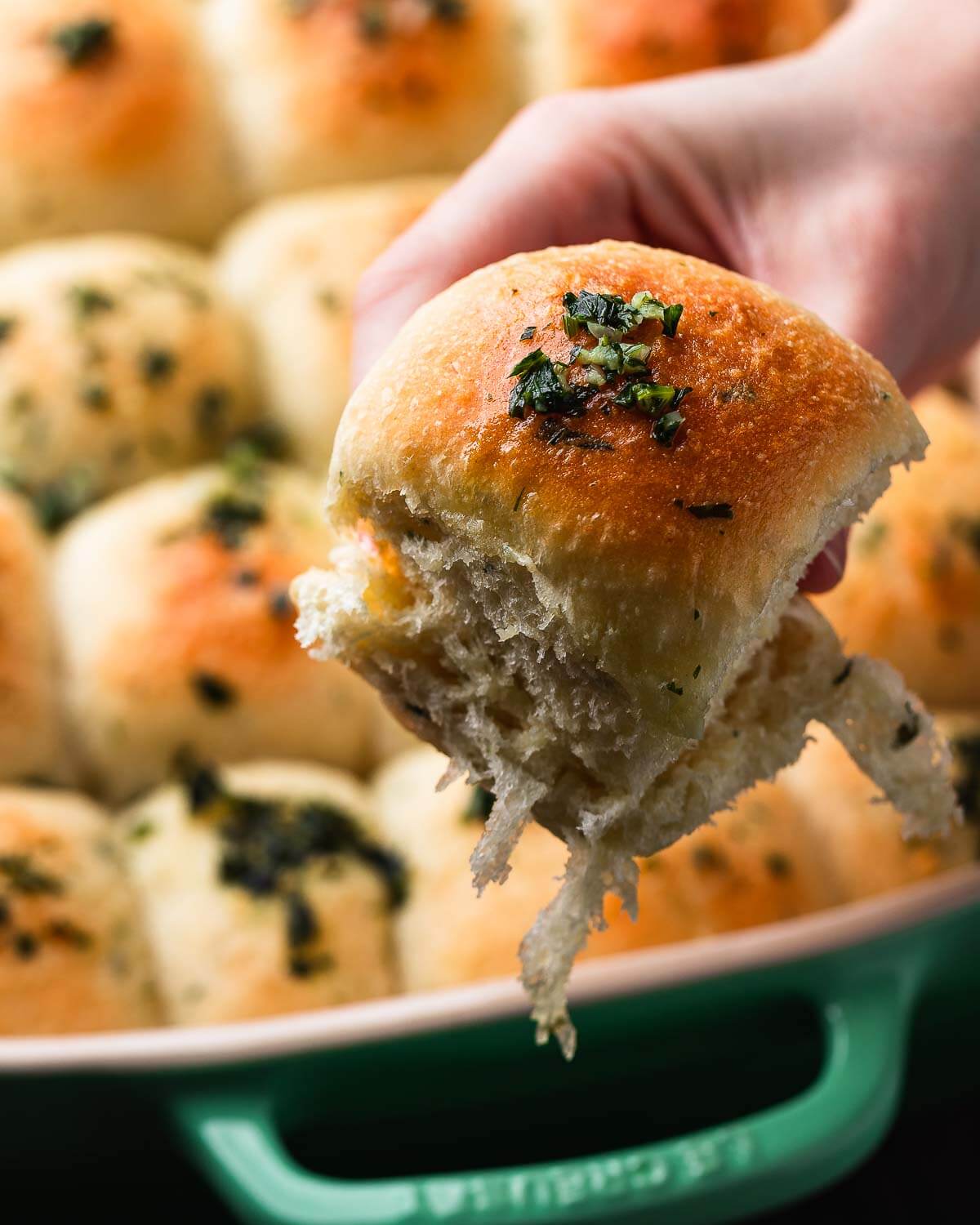 A hand holding a garlic butter roll over baking dish.