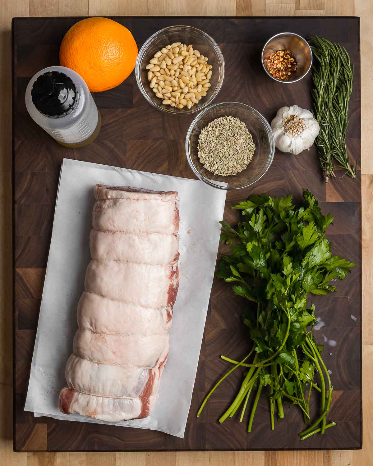 Ingredients shown: orange, olive oil. pine nuts, fennel seeds, hot red pepper flakes, garlic, rosemary, parsley, and tied pork loin.