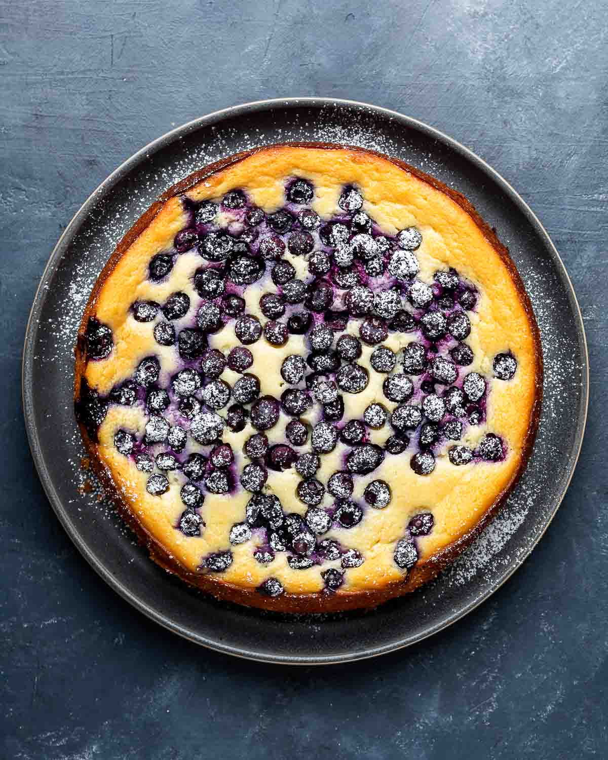 Overhead shot of lemon blueberry ricotta cake in grey plate on blue board.