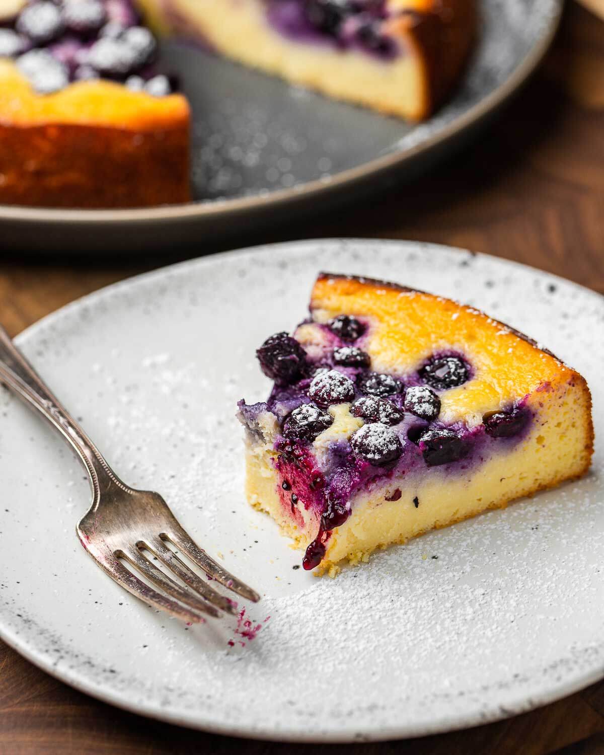 Bite taken out of slice of lemon blueberry ricotta cake on white plate.