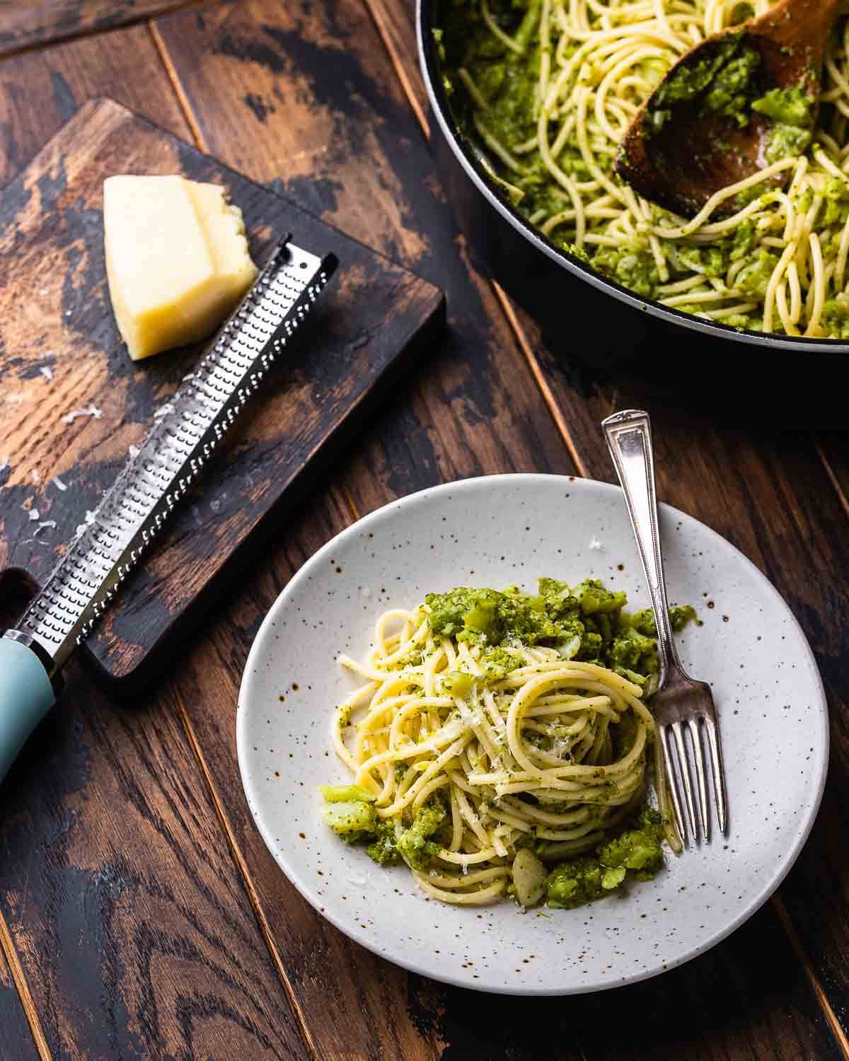 Table with white plate with spaghetti and broccoli along with a large pan and board with a block of Pecorino Romano cheese.