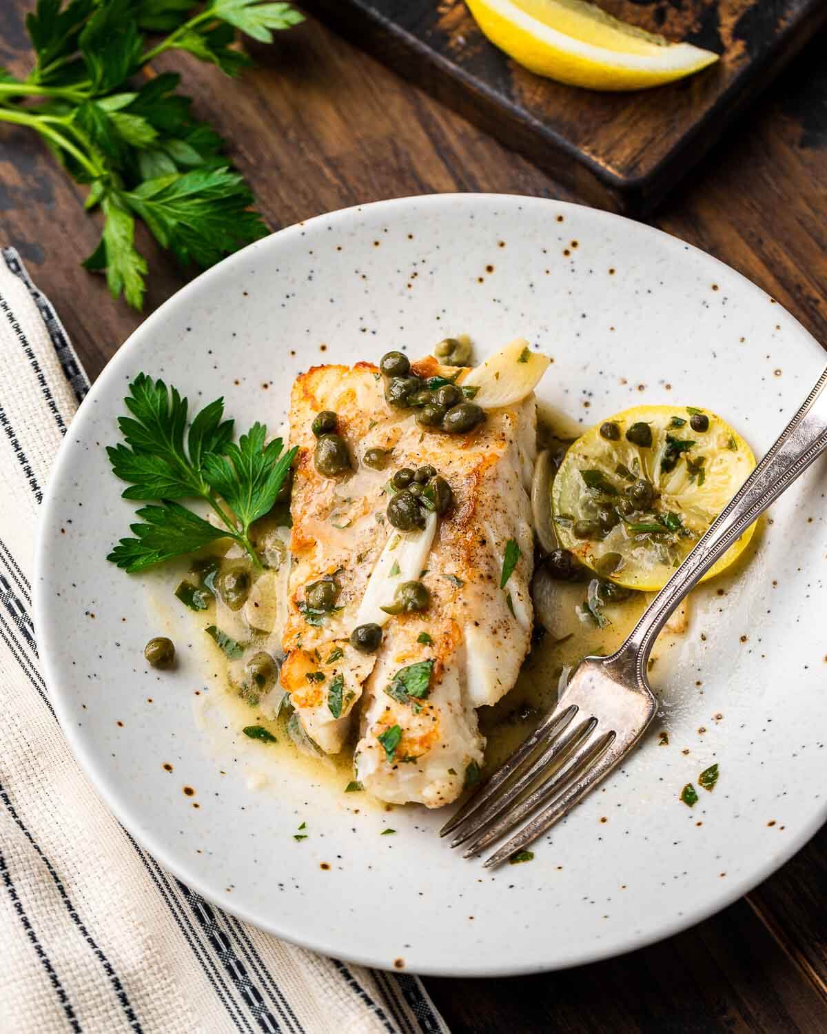 Cod piccata in white plate on brown table with parsley and lemon wedges in the background.