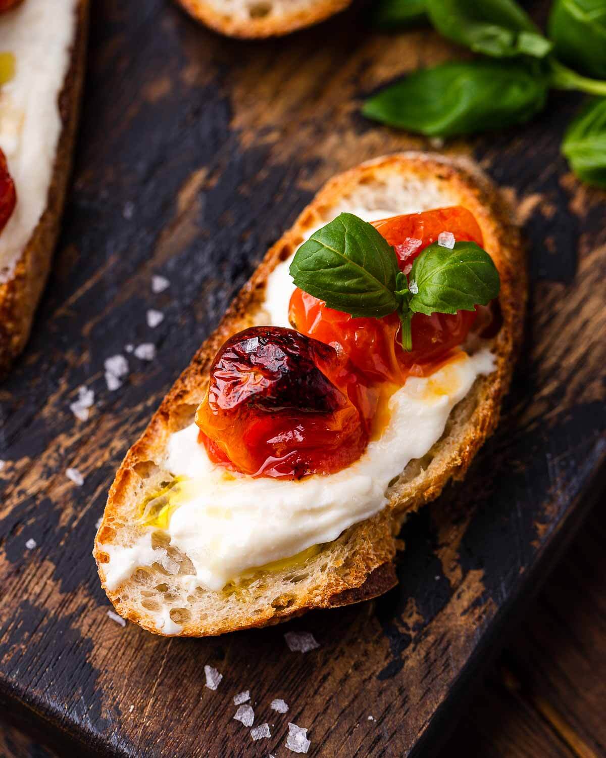 Ricotta roasted cherry tomato crostini with basil and flaky sea salt on wooden board.