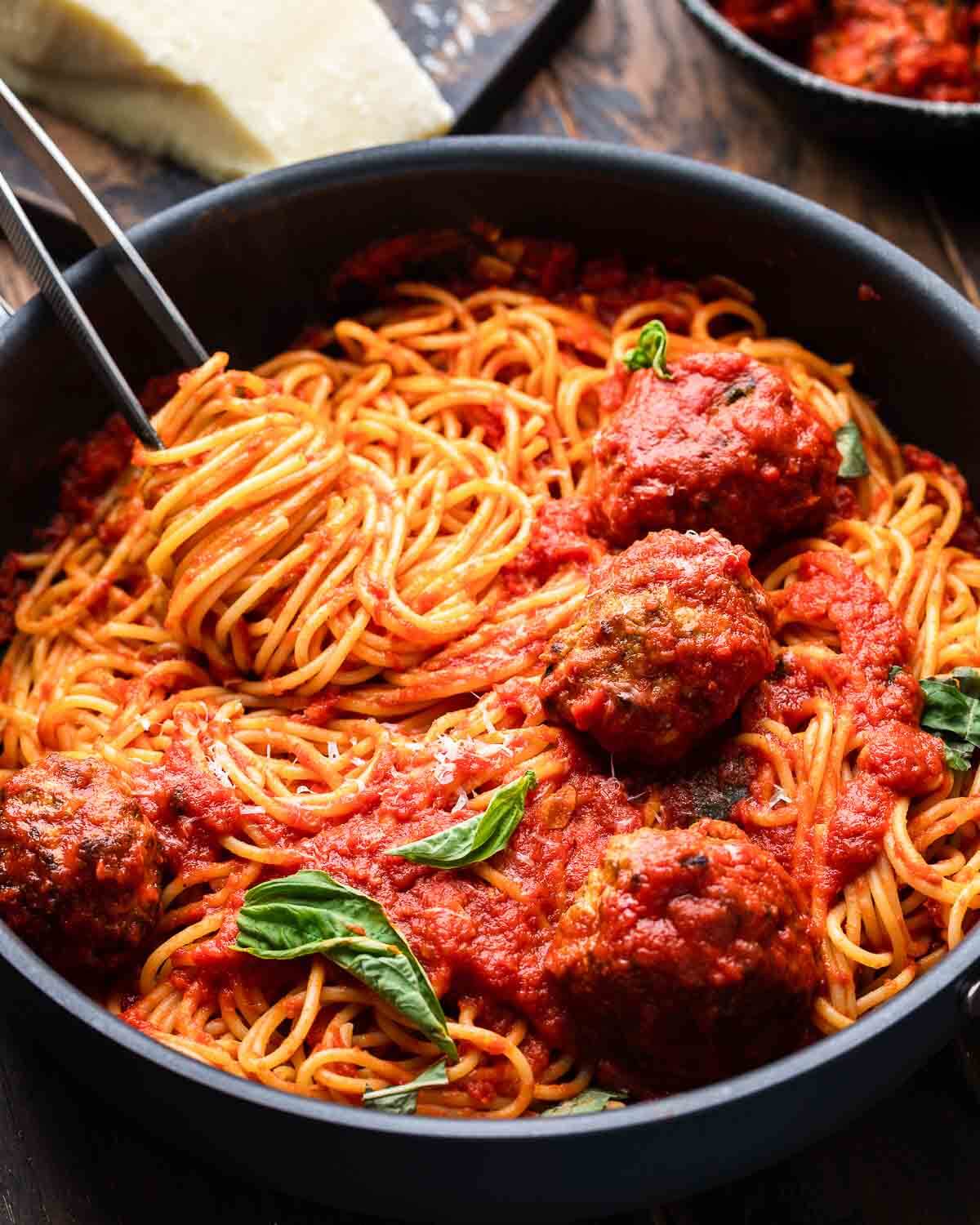 Spaghetti and meatballs in black pan with block of cheese and bowl of meatballs in the background.