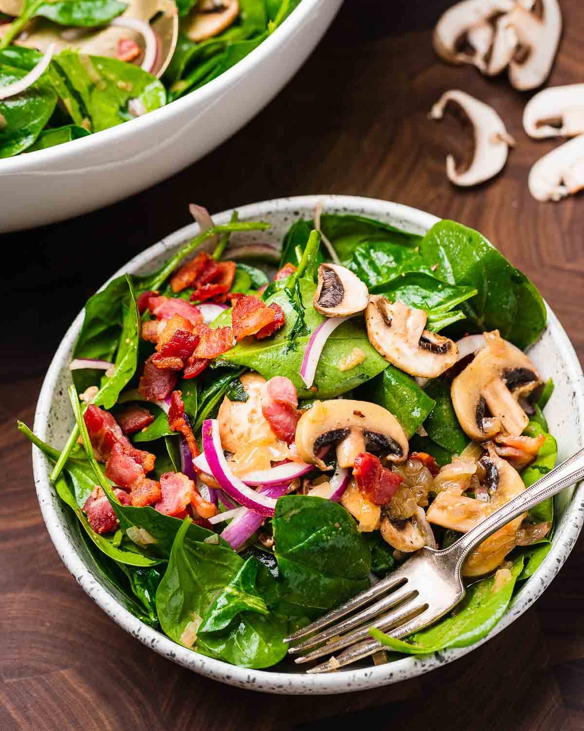 Spinach salad with hot bacon dressing in white bowl with mushrooms on the table.