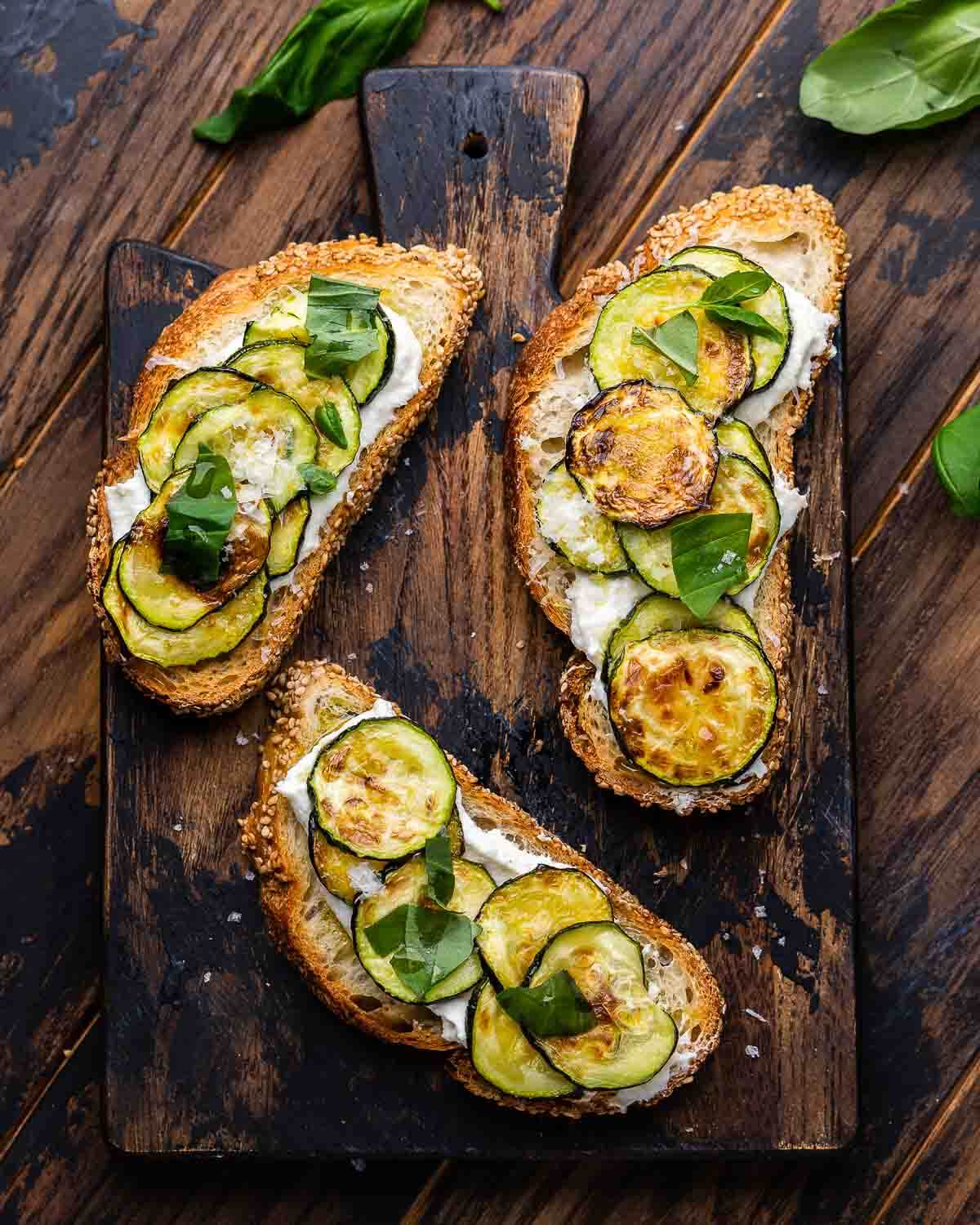 Overhead shot of zucchini bruschetta on wooden board with basil leaves spread around it.