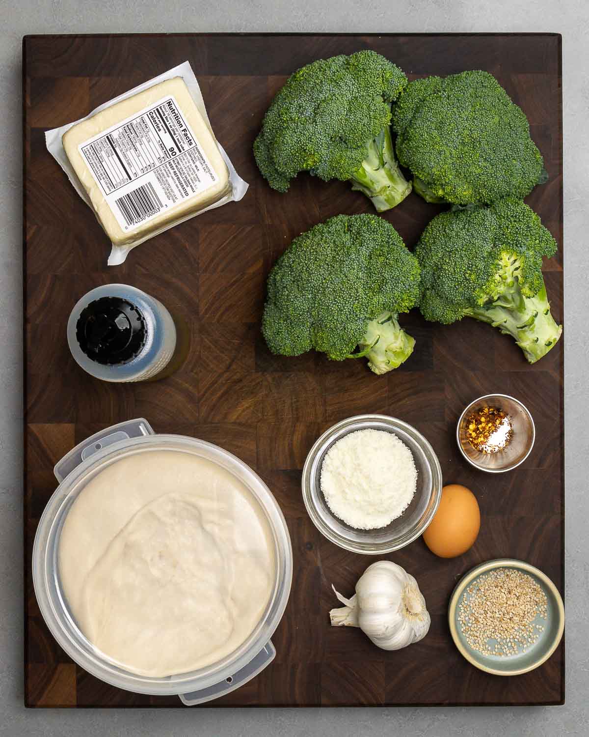 Ingredients shown: mozzarella, broccoli crowns, olive oil, Pecorino Romano, eggs, hot red pepper flakes, garlic, sesame seeds, and pizza dough.