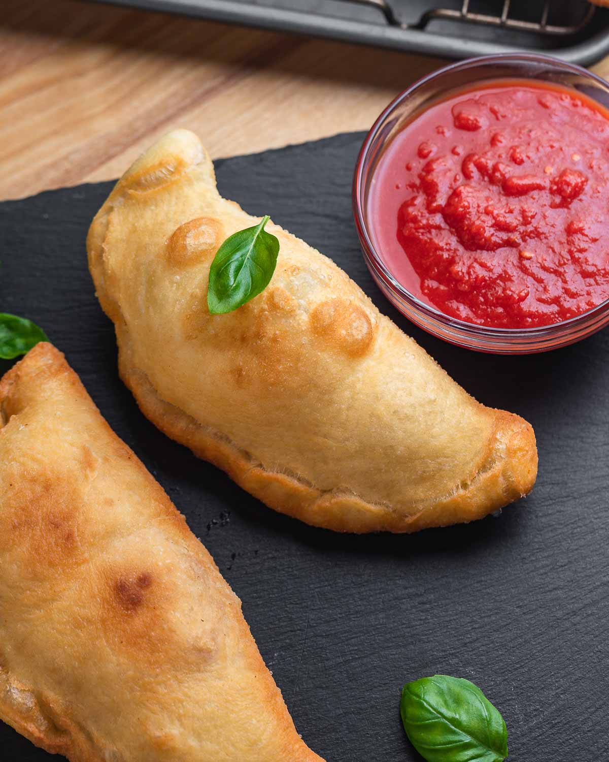 Two panzerotti on black slate with basil leaves and a bowl of sauce.