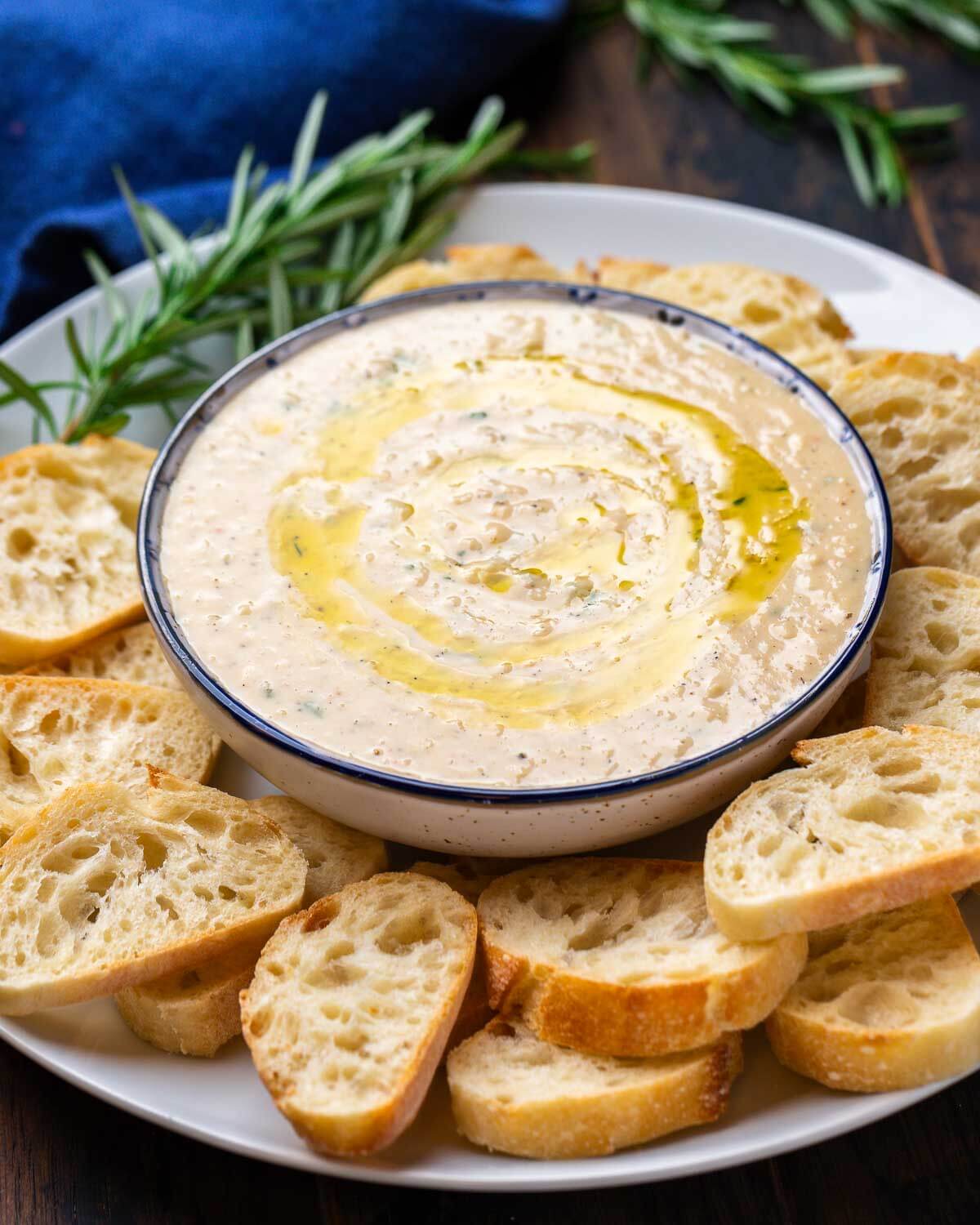 Bowl of cannellini bean dip with crostini and rosemary garnish.