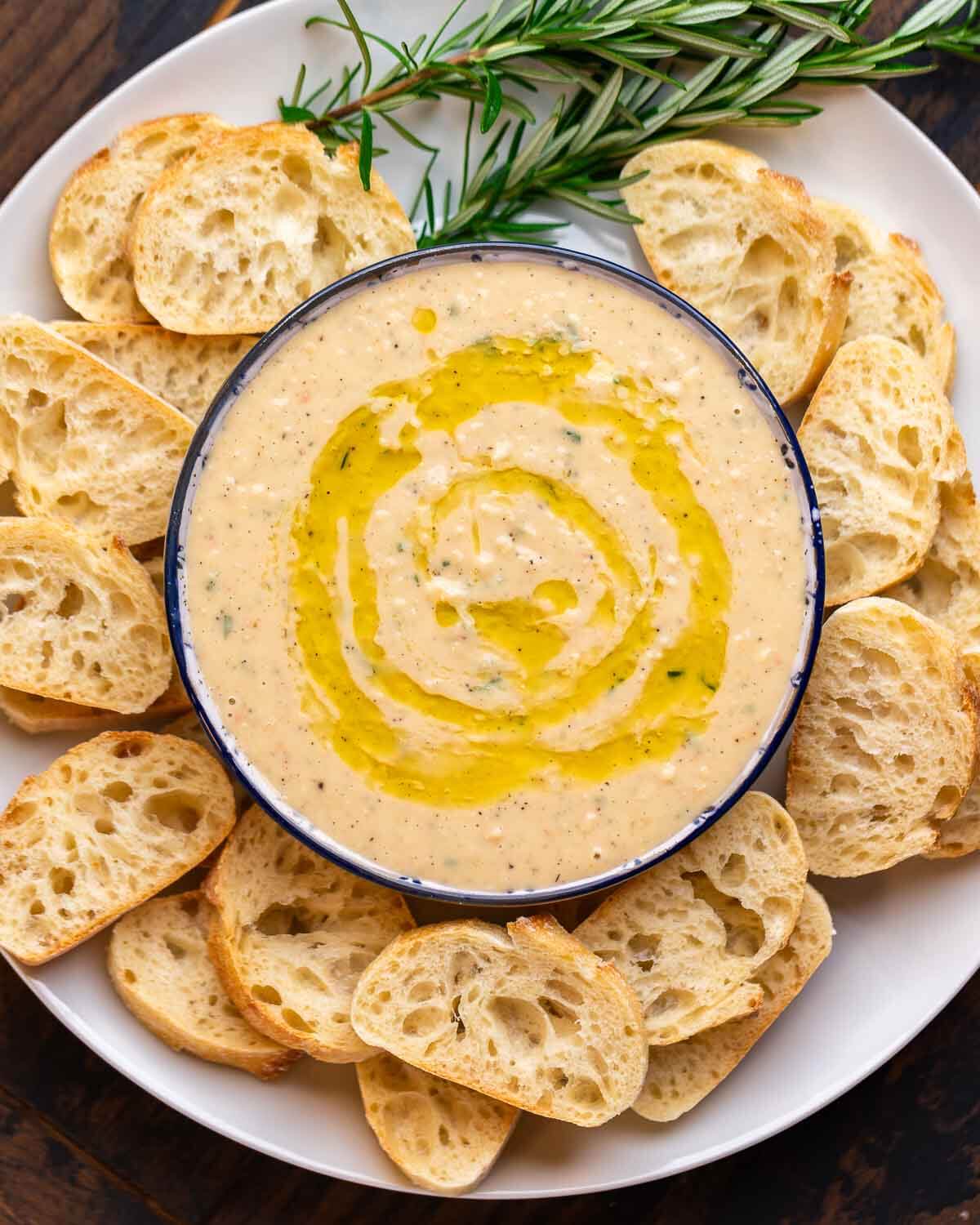 Overhead shot of cannellini bean dip platter with crostini and rosemary garnish.
