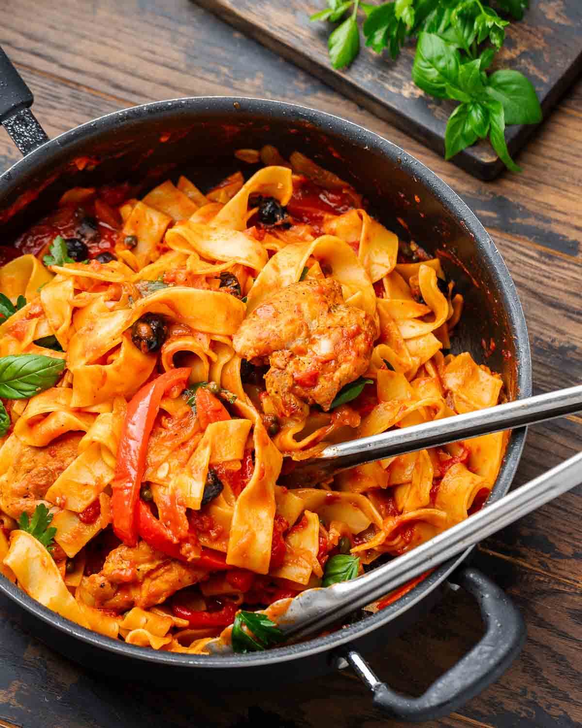 Large pan with chicken cacciatore pasta with tongs and board with basil and parsley.