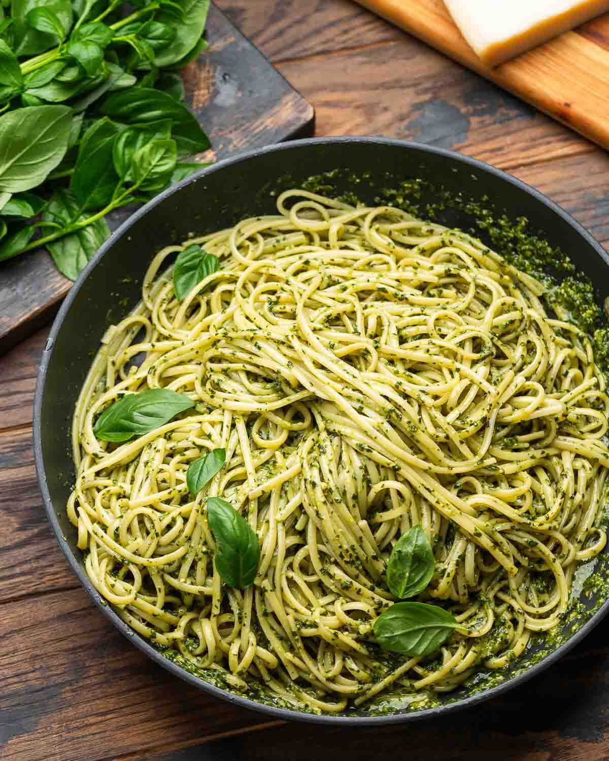 Large black pan with pasta with pesto Genovese along with basil and cheese in the background.