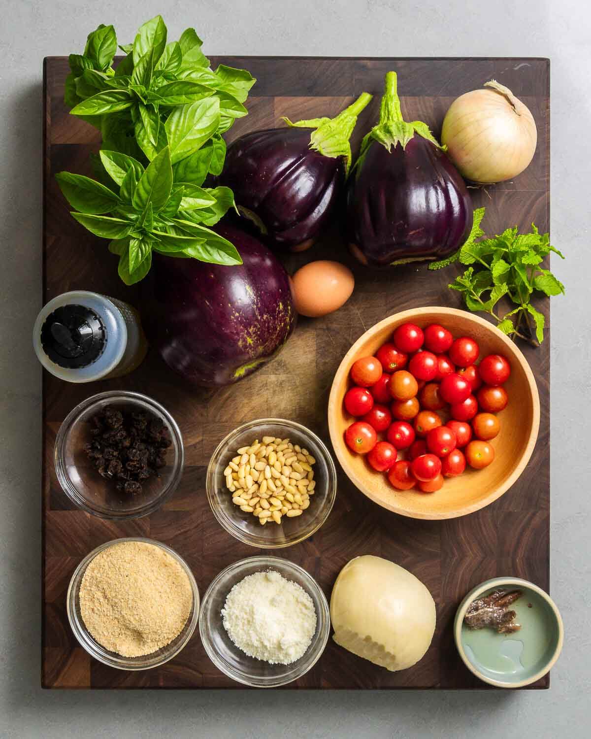 Ingredients shown: basil, mint, eggplant, onion, garlic, egg, olive oil, raisins, pine nuts, cherry tomatoes, breadcrumbs, Pecorino, Scamorza, and and anchovies.