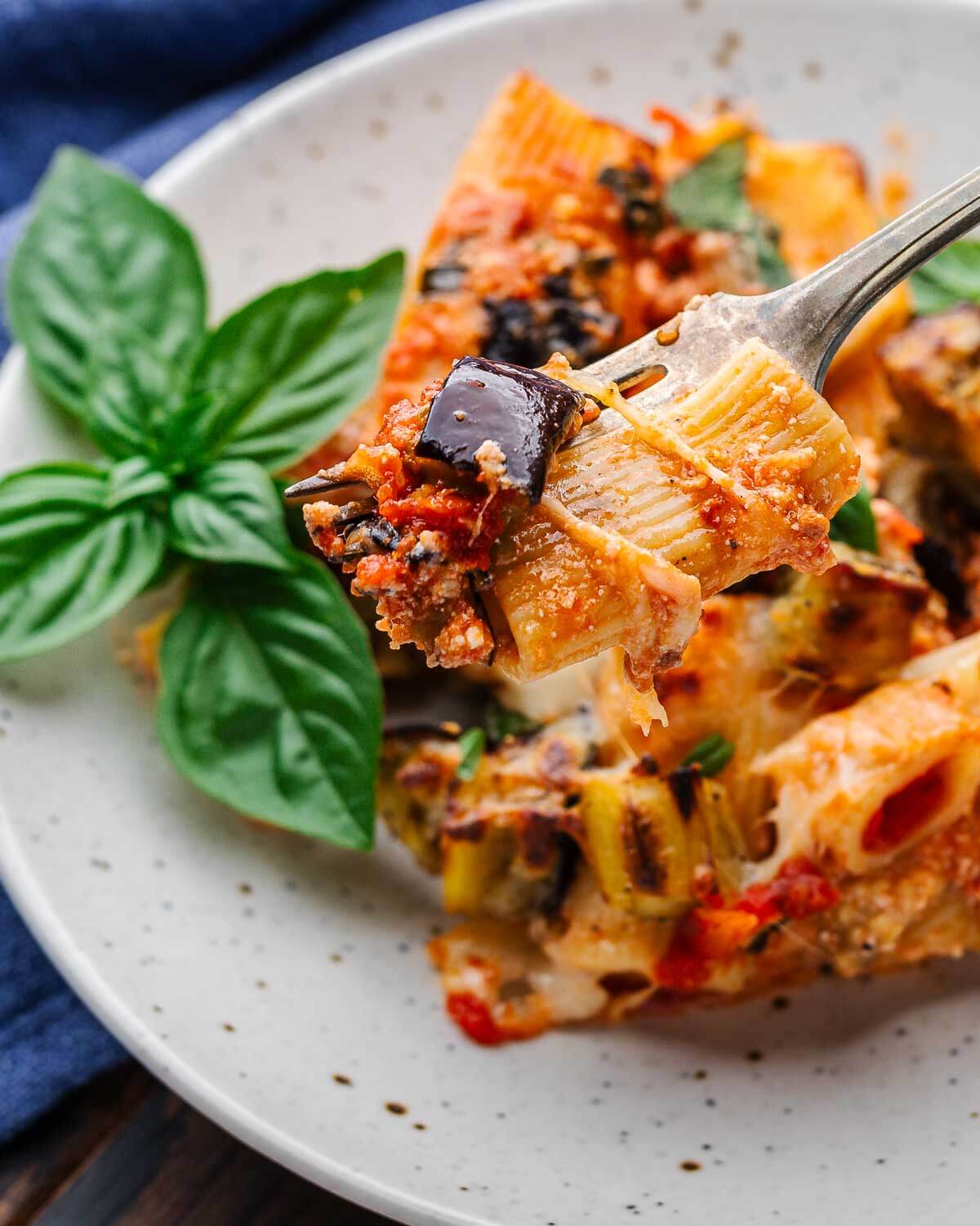 Fork holding baked rigatoni with eggplant over white plate.