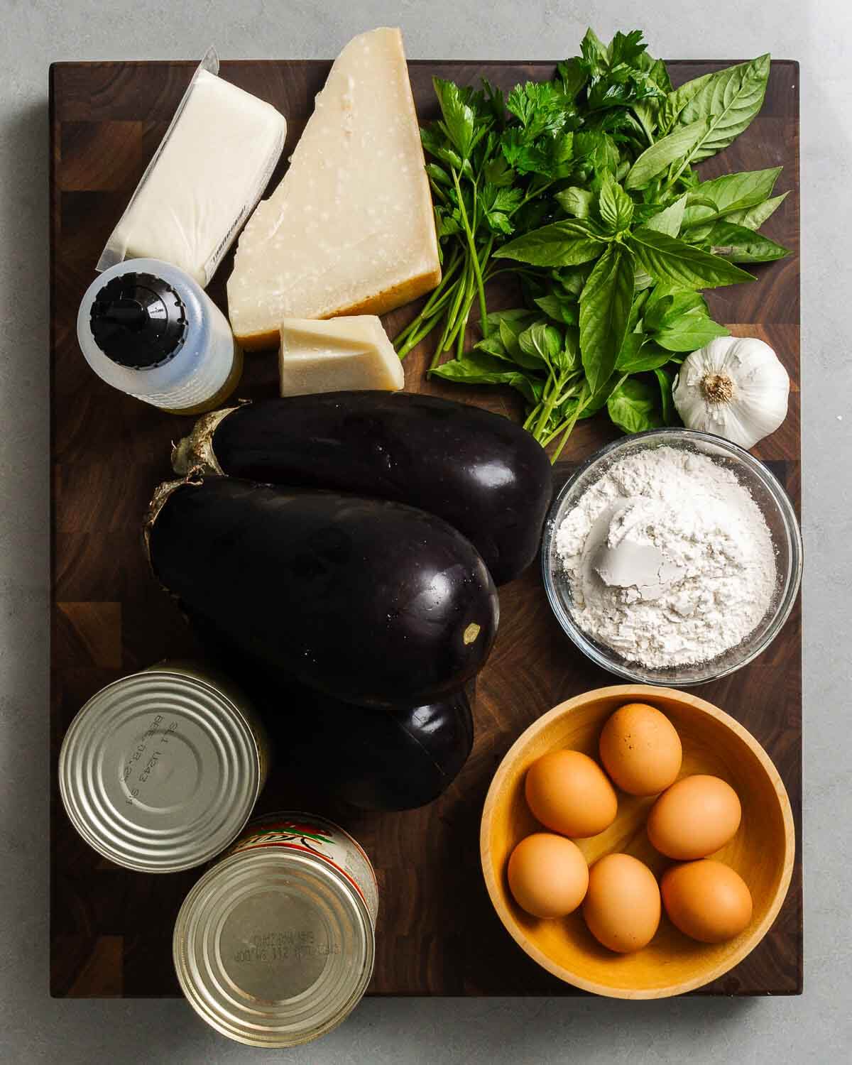 Ingredients shown: olive oil, mozzarella, parmesan, pecorino, parsley, basil, garlic, eggplants, flour, tomatoes, and eggs.