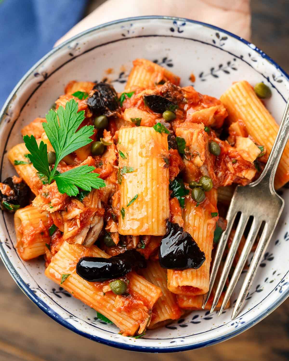 Hands holding small bowl of pasta al tonno with fork and parsley garnish.