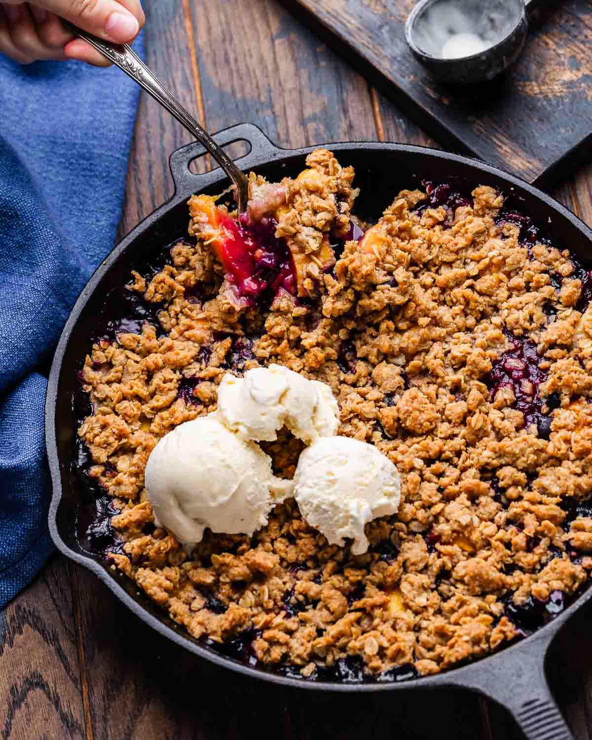 Peach blueberry crisp in cast iron pan on table with spoon scooping out some of it.