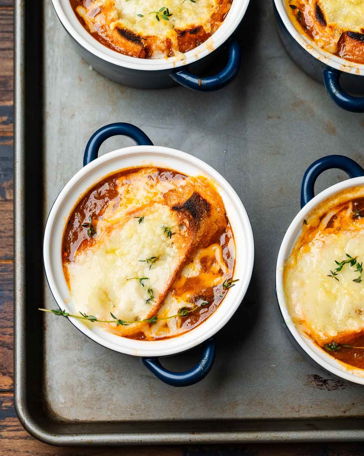 Overhead shot of four blue crocks with baked French onion soup on baking sheet.