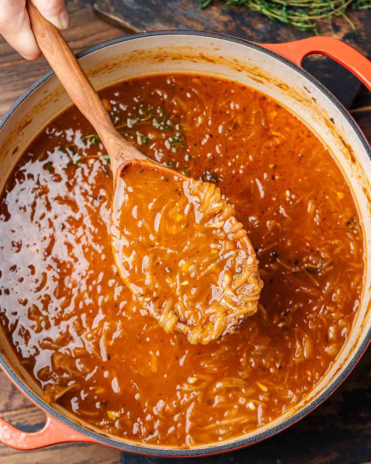 Hands holding wooden ladle with French onion soup over large Dutch oven.