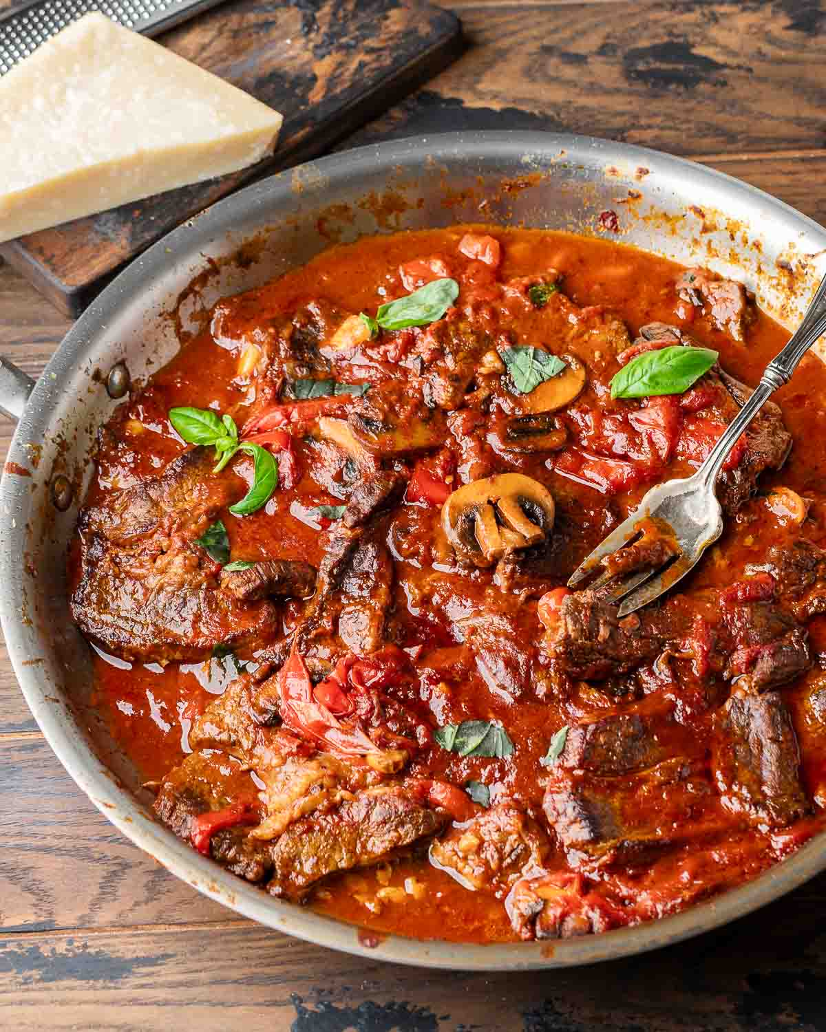 Pan with steak pizzaiola and block of parmesan cheese on wood table.