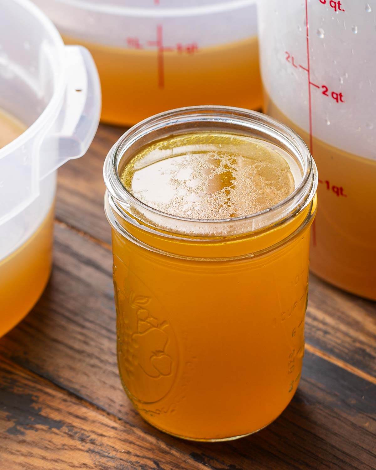 Mason jar filled with beef stock along with 3 large containers in the background.