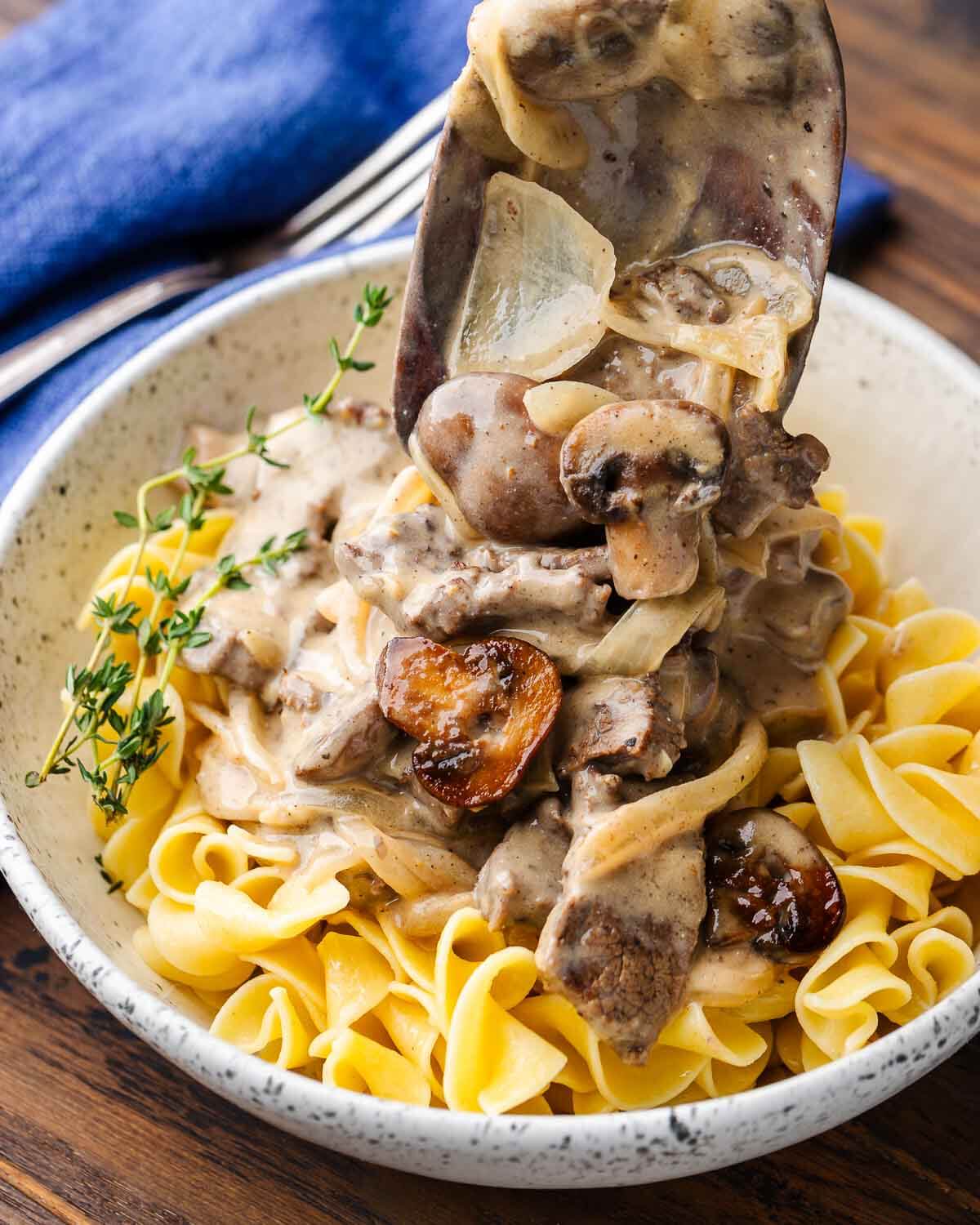 Wooden spoon placing beef stroganoff on egg noodles in white bowl.