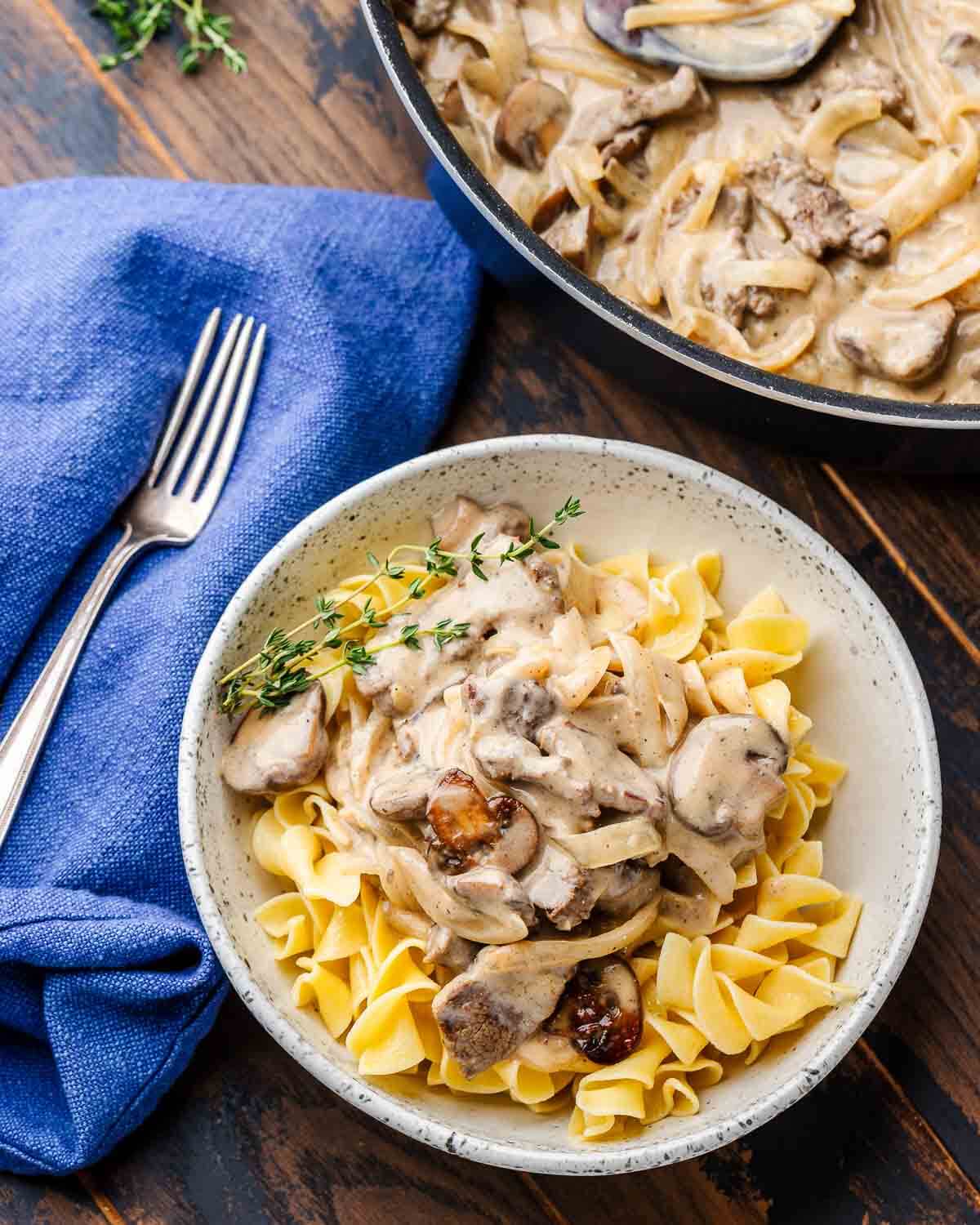 White bowl with beef stroganoff, blue napkin, and fork.