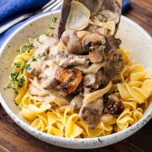 Beef stroganoff featured image with wooden spoon pouring gravy over egg noodles in white bowl.