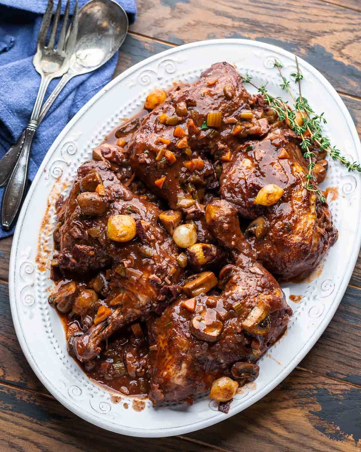 Overhead shot of coq au vin in white serving platter on wood table.