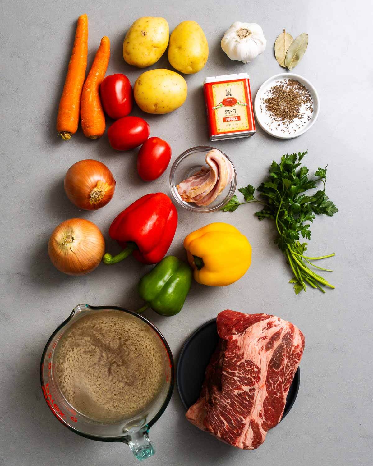 Ingredients shown: carrots, potatoes, garlic, bay leaves, Hungarian paprika, caraway seeds, plum tomatoes, bacon, onion, peppers, parsley, beef stock, and chuck roast.