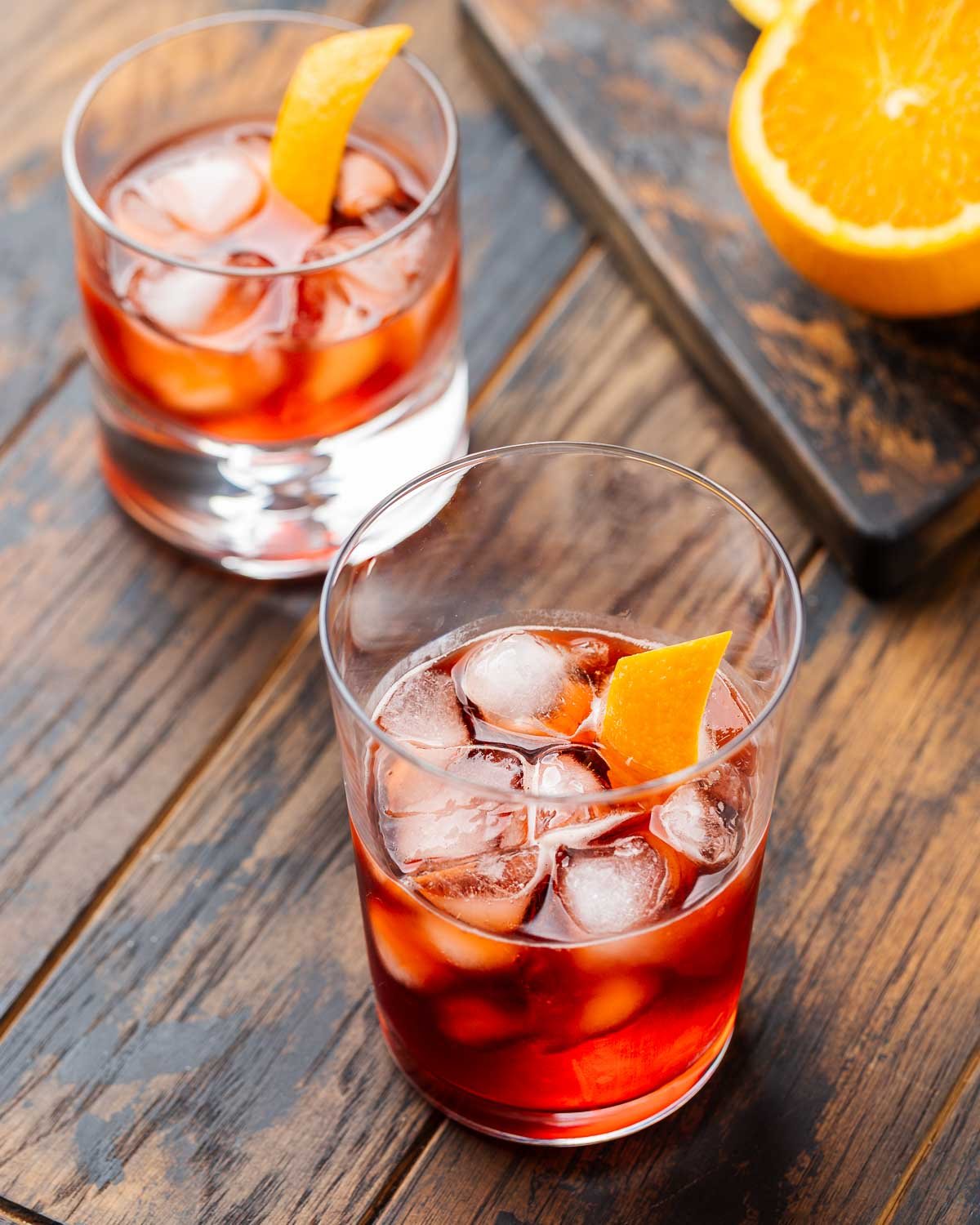 Two Negronis on a wooden board with a cut orange in the background.