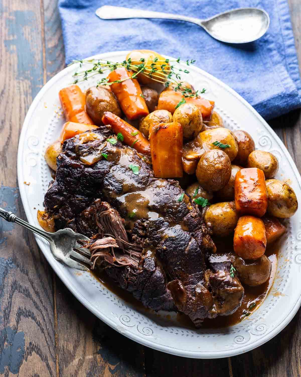 Large white platter with pot roast, carrots, potatoes, and blue napkin and serving spoon on the side.