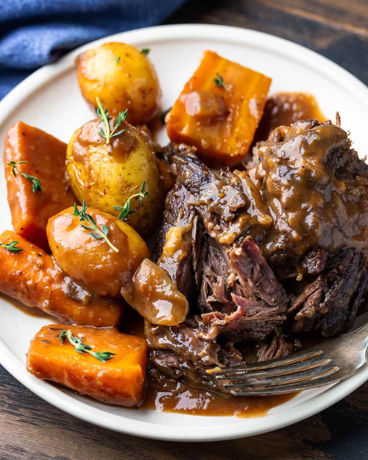 White plate with vegetables and piece of pot roast torn open by fork.