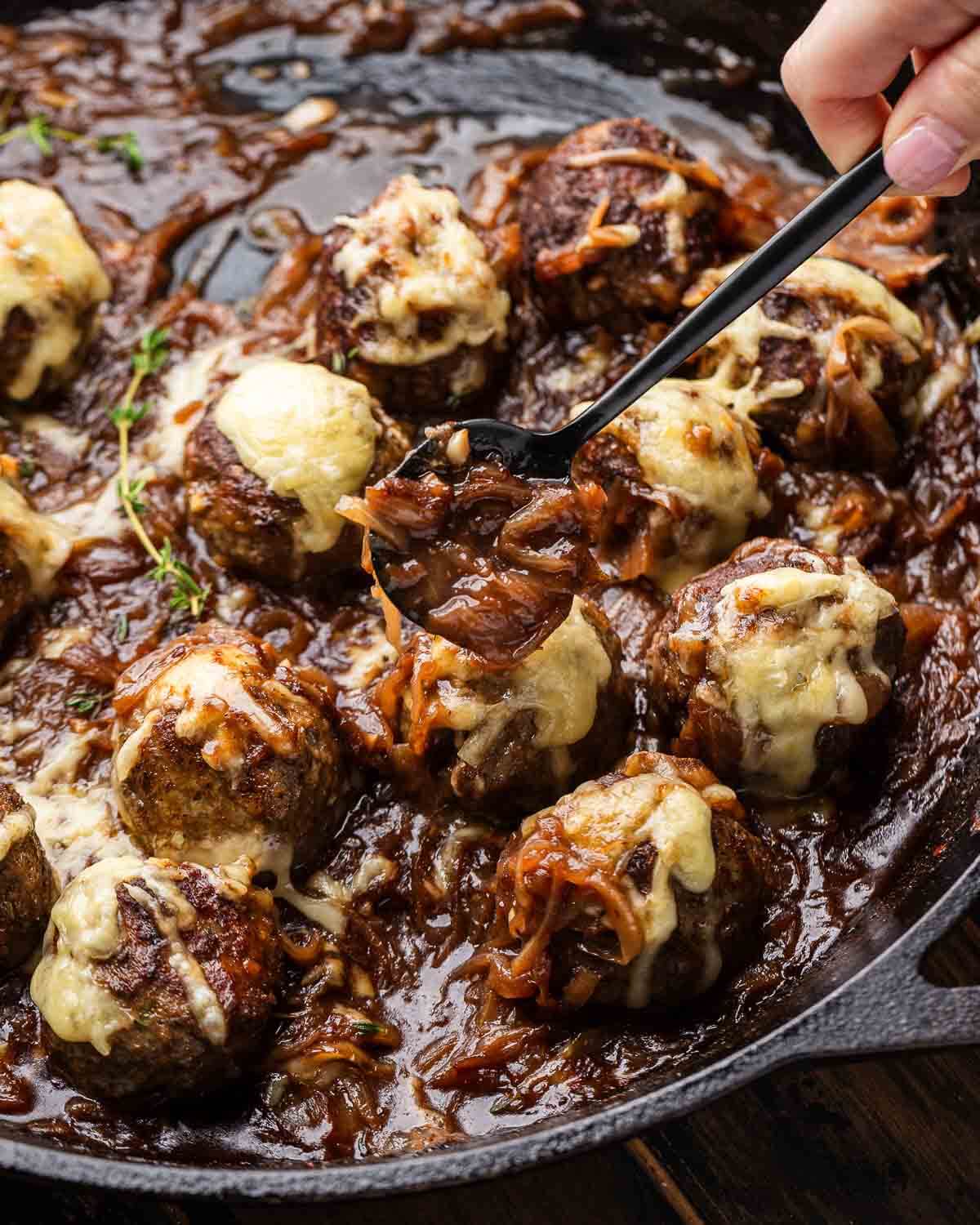 Hands holding spoonful of onions above meatballs in cast iron pan.
