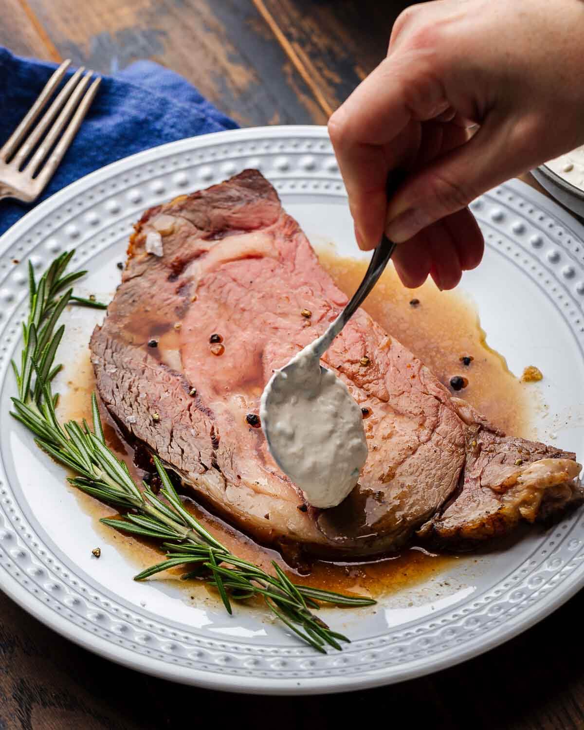 Hands spooning horseradish sauce onto a slice of prime rib in white plate.