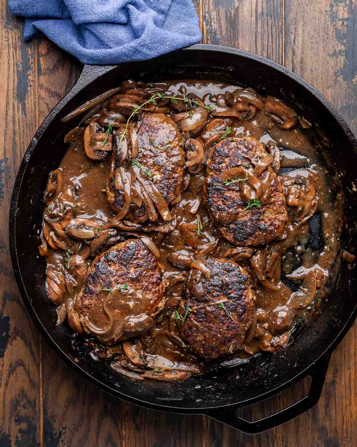 Cast iron pan with salisbury steaks topped with mushroom gravy on wood table.