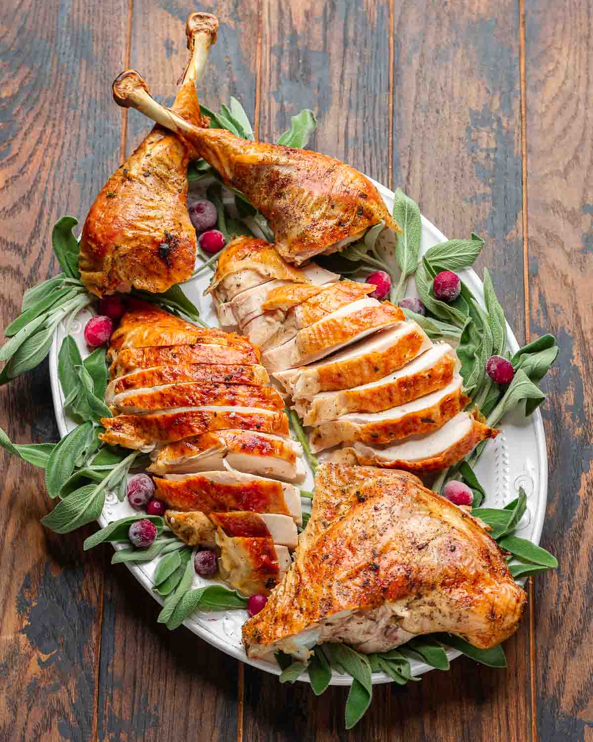 Whole roast turkey sliced on white platter with sage and cranberry garnish.