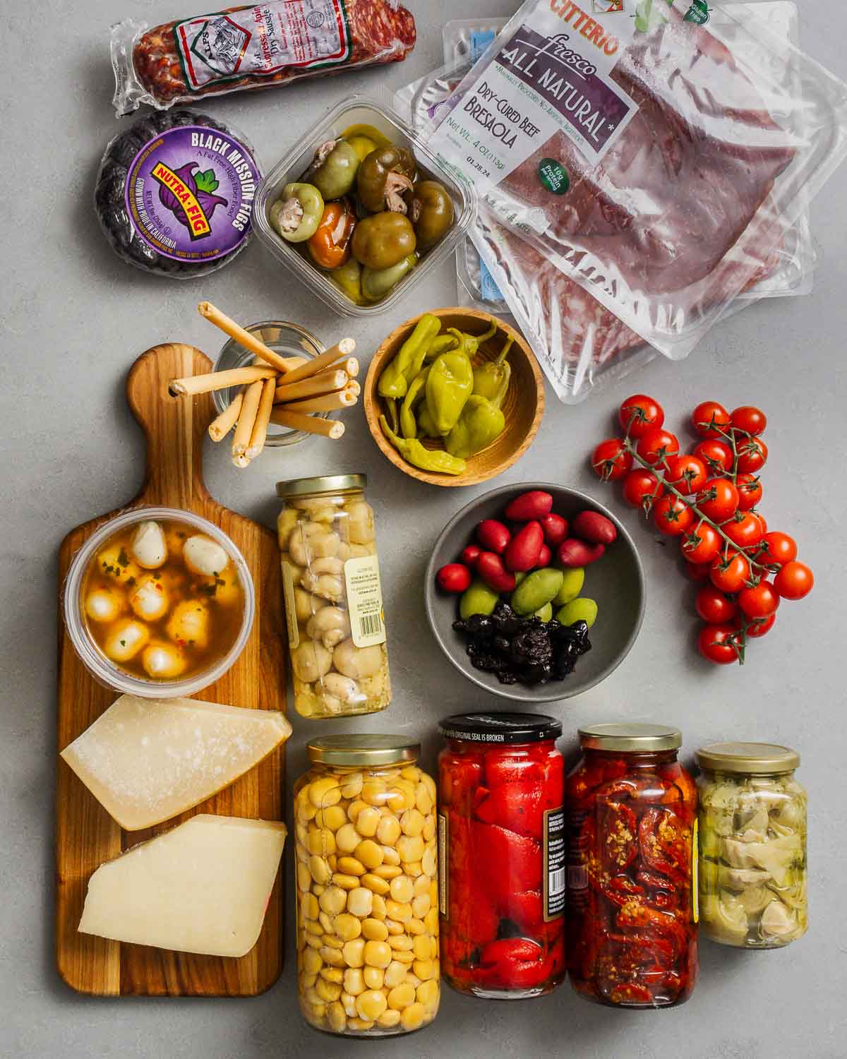 Ingredients shown: salami, figs, stuffed cherry peppers, cured meats, breadsticks, mozzarella balls and assorted cheeses, pickled mushrooms, red roasted peppers, sun-dried tomatoes, artichoke hearts, olives, and cherry tomatoes.