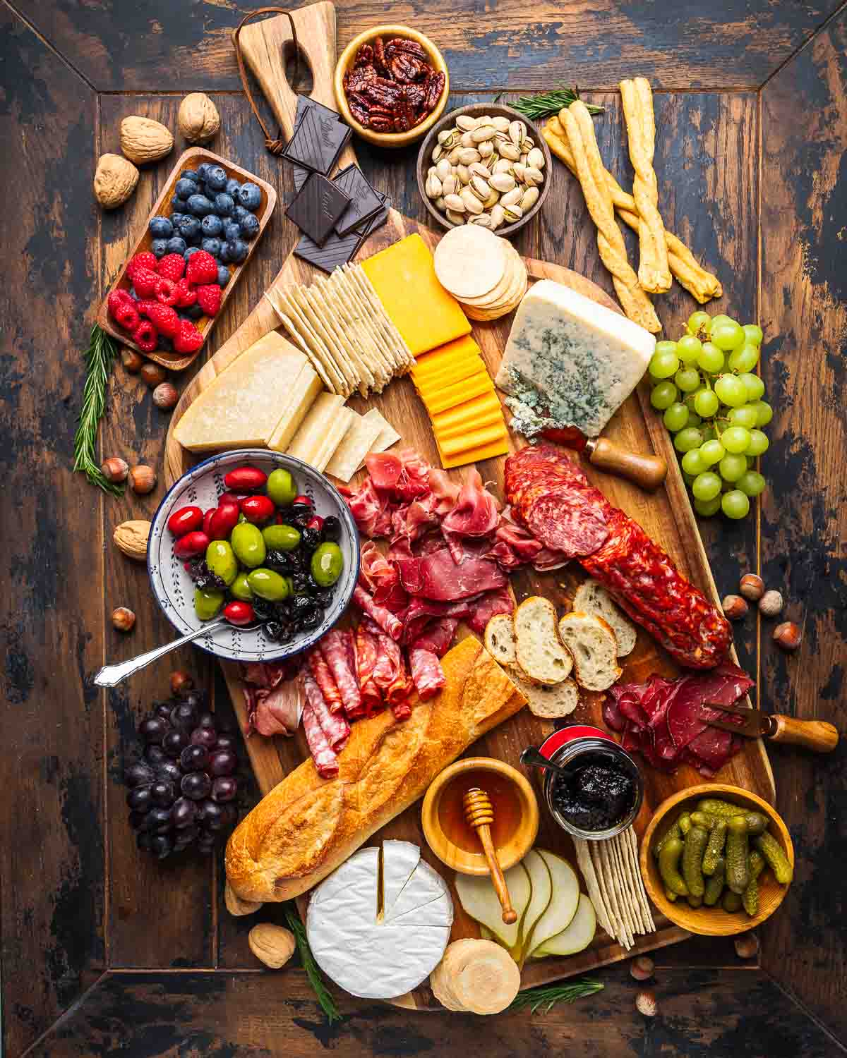 Overhead shot of massive charcuterie board with cheeses, cold cuts, olives, honey, jam, grapes, nuts, chocolate, and baguette.