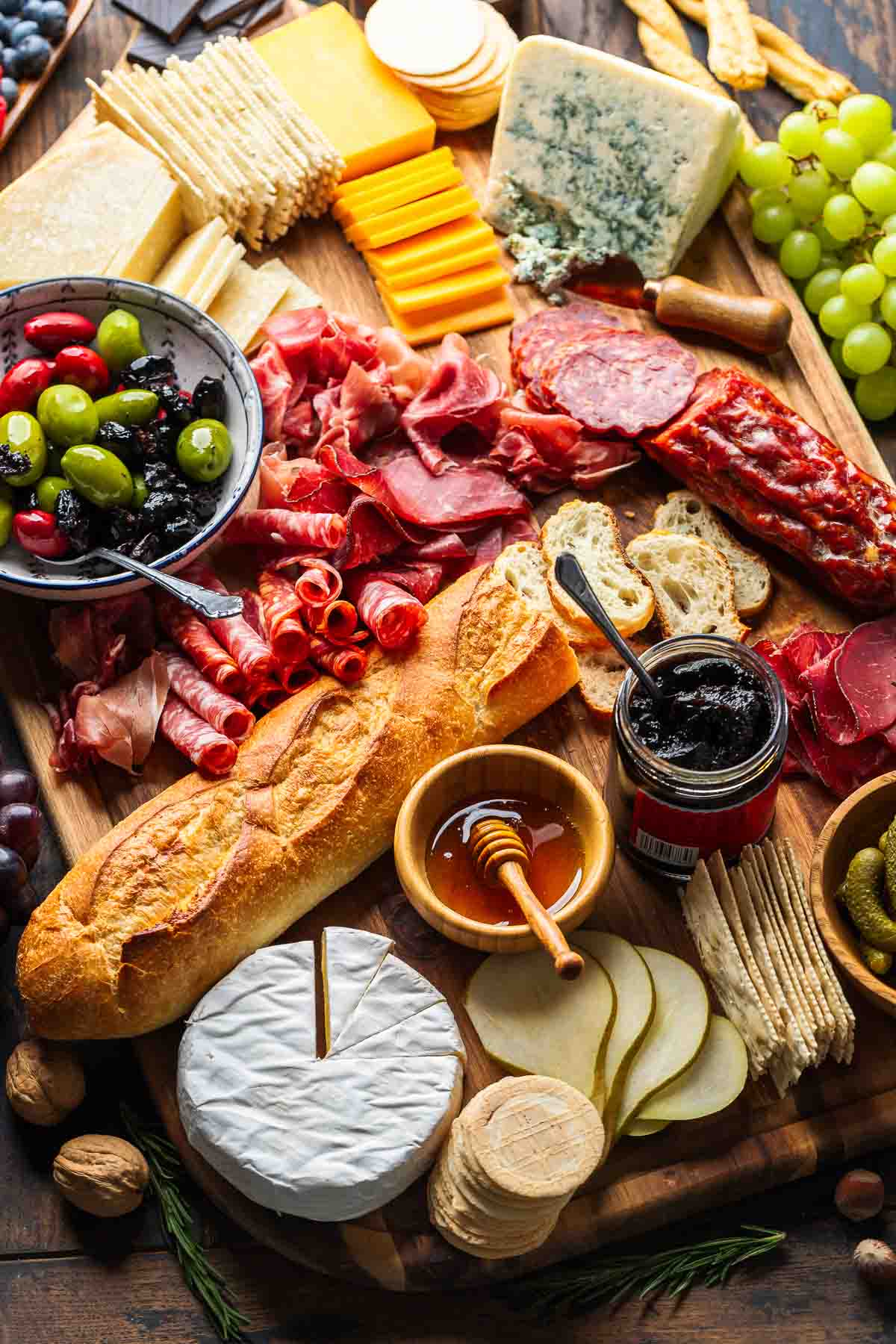 Charcuterie board with cheeses, cold cuts, olives, honey, jam, grapes, and baguette.