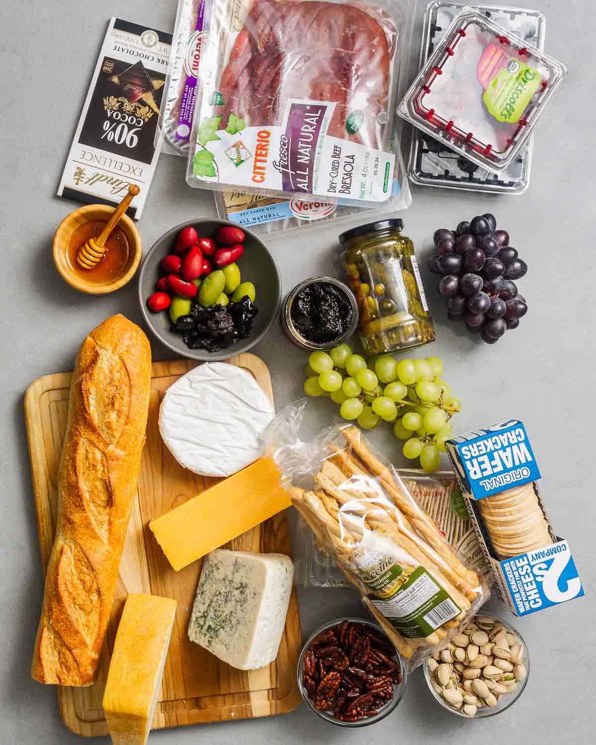 Ingredients shown: chocolate, cured meats, berries, honey, olives, jam, cornichon, grapes, baguette, cheeses, breadsticks, nuts, and crackers.
