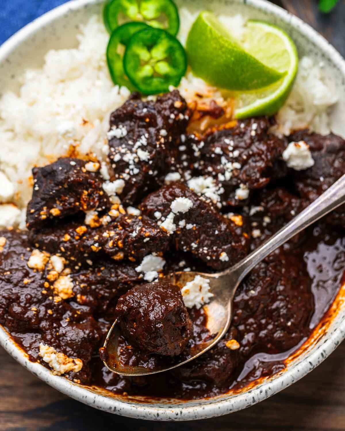 White bowl with chili colorado, white rice, and garnished with limes, cilantro, and cotija cheese.