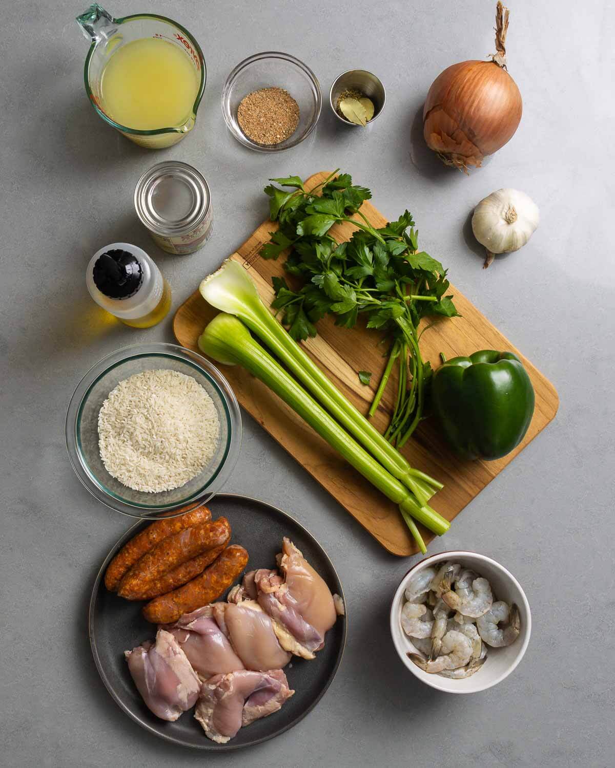 Ingredients shown: chicken stock, Cajun seasoning, spices, onion, garlic, tomato sauce, olive oil, celery, bell pepper, parsley, rice, Andouille sausage, chicken thighs, and shrimp.