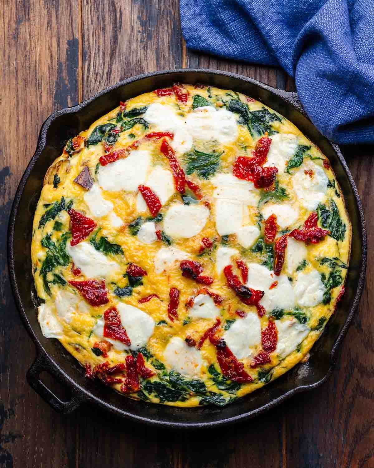 Overhead shot of sun-dried tomato frittata in cast iron pan with blue kitchen towel.