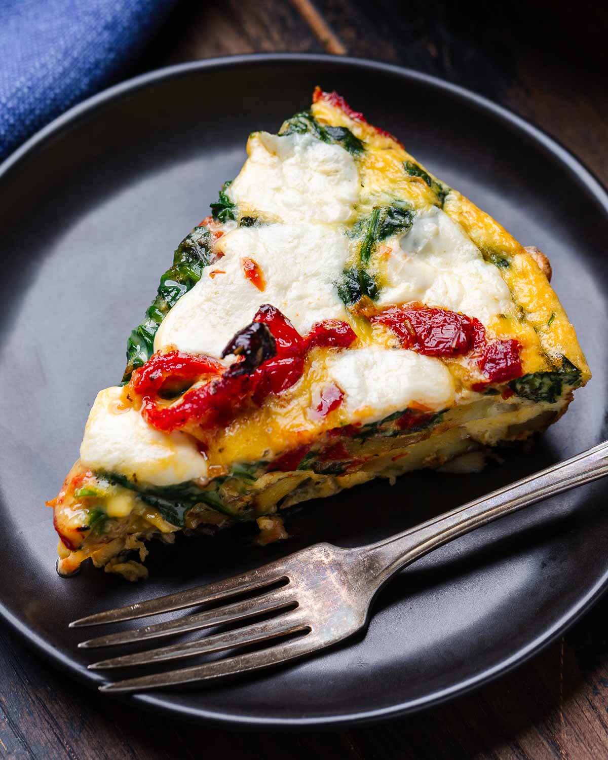 Black plate with slice of sun-dried tomato, spinach, and fresh mozzarella frittata.