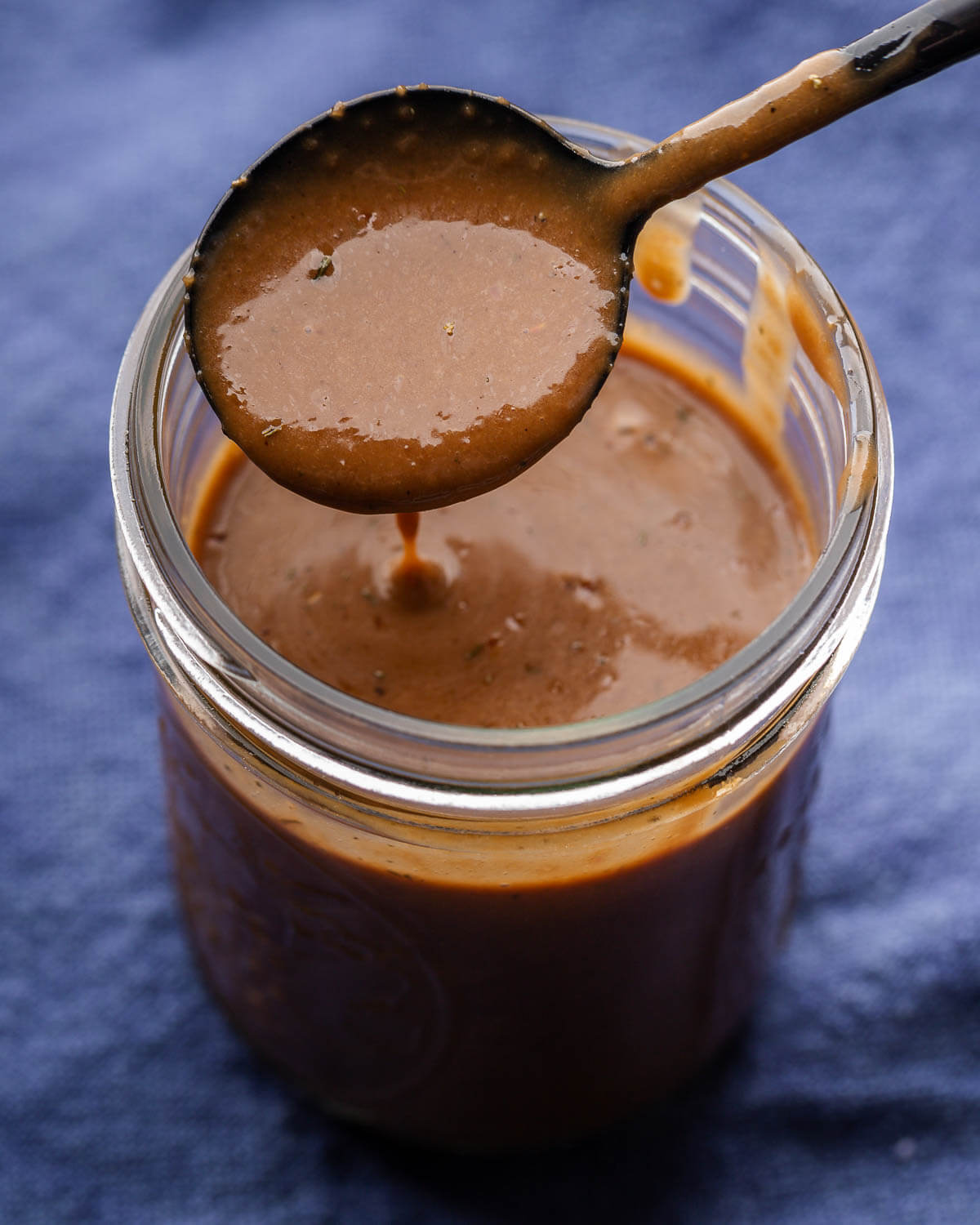 Black spoon drizzling balsamic vinaigrette over mason jar on blue napkin.